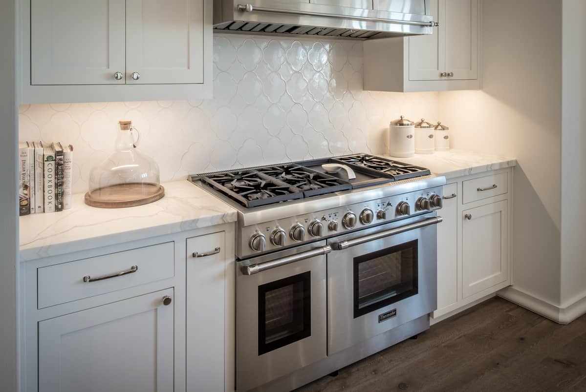 A stainless steel oven set into white cabinetry. There are white marble countertops and white tile backsplash.