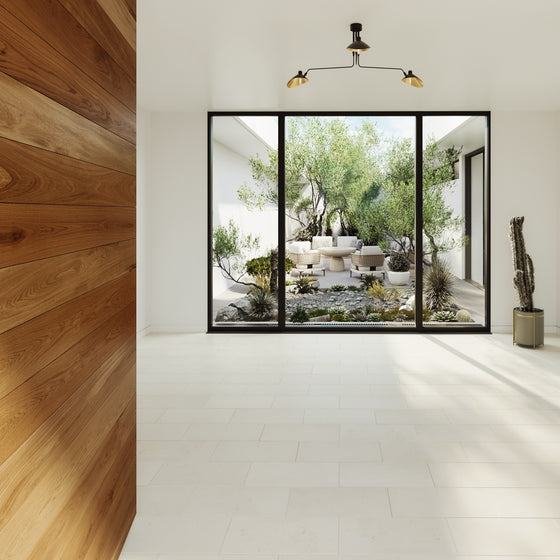 Modern interior with wooden wall paneling, a white tile floor, and large windows showcasing a lush garden with seating, rocks, and plants. Soft lighting above.