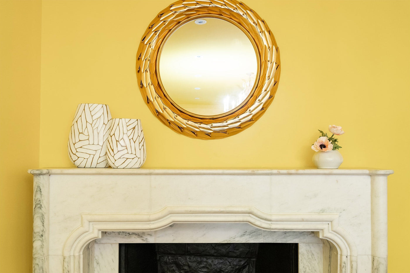 A white marble fireplace against a yellow wall. The fireplace has several decorative vases sitting on top of it, and there is a circular gold mirror above it.