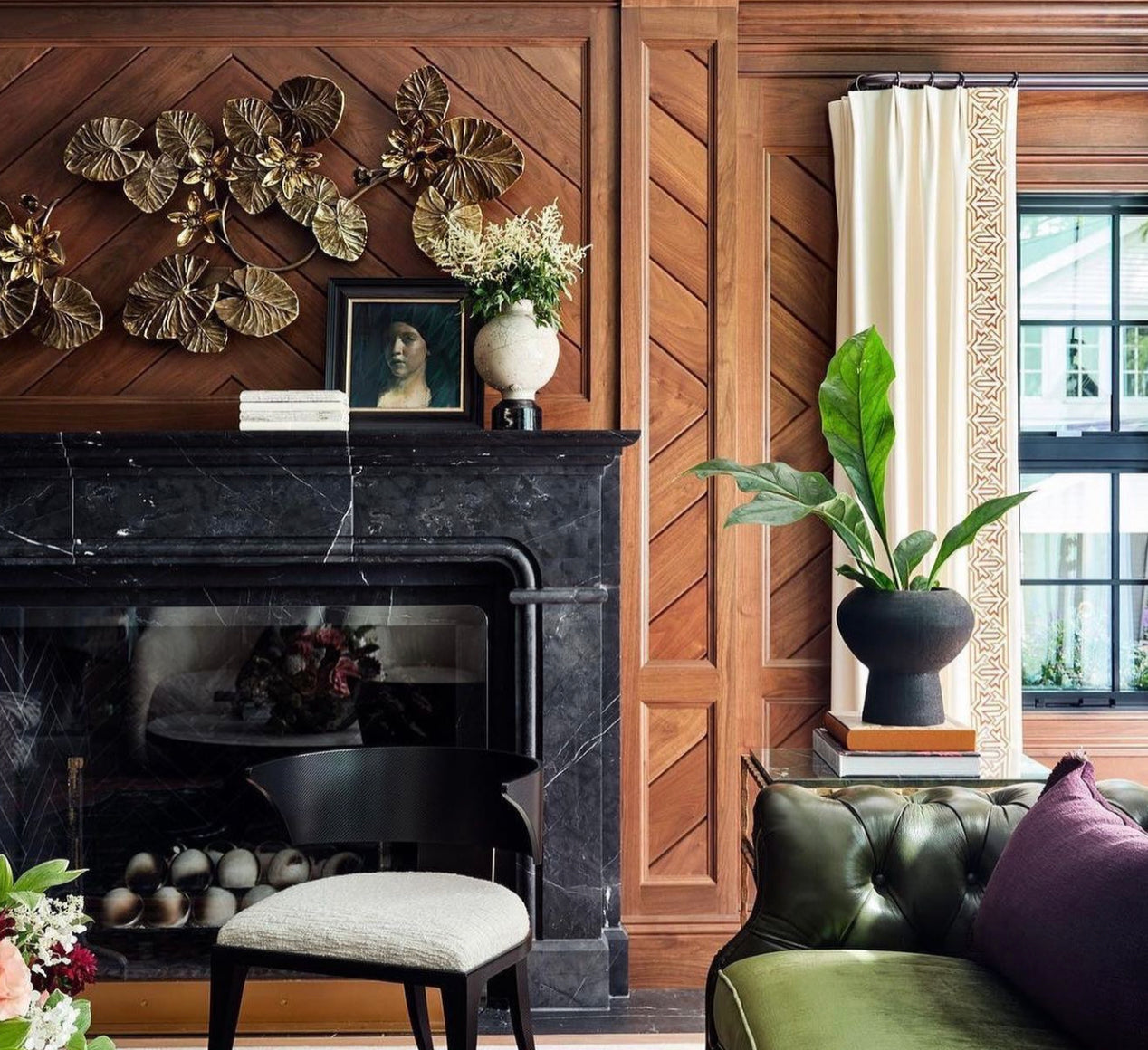 black marble fireplace in a wood-paneled living room. There is a green couch and a black chair with a white seat in the foregroundnd.