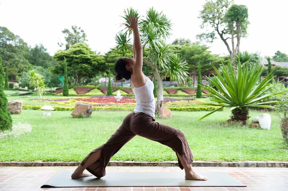 A woman practices yoga outside. There is a grass yard with various trees and decorative plants behind her.