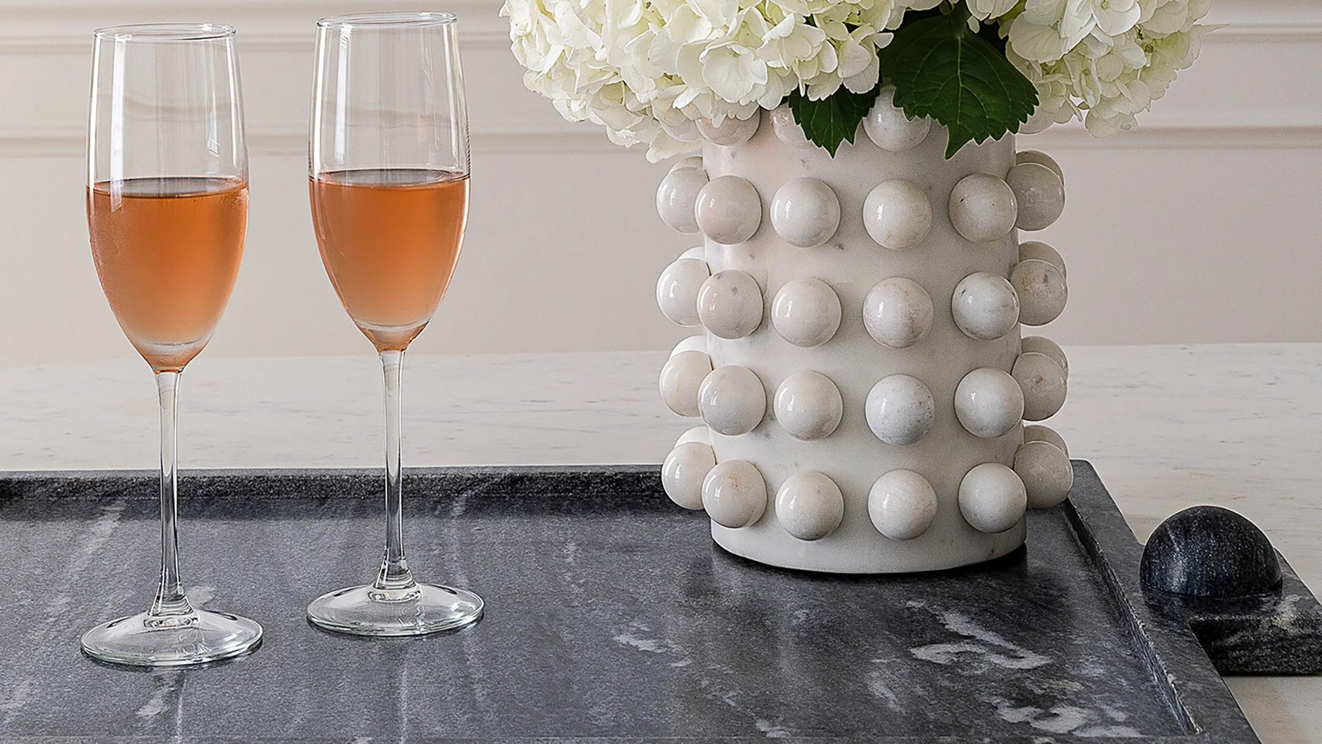 Two glasses of rose wine sit on a dark marble tray beside a textured white marble vase filled with white hydrangeas.