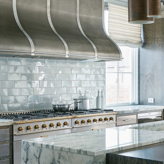 Sleek kitchen with a metallic range hood, white tiled backsplash, and marble countertops. Pots sit on a modern stove, evoking a clean, elegant vibe. - Mobile View