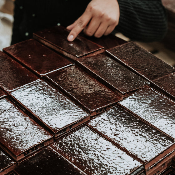 A hand wearing a black sleeve points at a stack of shiny, rectangular, warm brown colored tiles arranged neatly.
